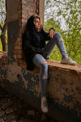 A beautiful woman with curly brown hair in rock Gothic grunge style: torn jeans, black mantle with hood, sitting on the broken window of an abandoned industrial building with brick walls. Loneliness