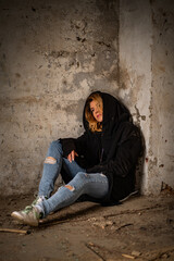 A mysterious goth woman in a black robe with a hood, torn jeans, sits thoughtfully on the dirty floor in an abandoned and destroyed building. Mysticism, post-apocalypse, depressed loneliness, witch