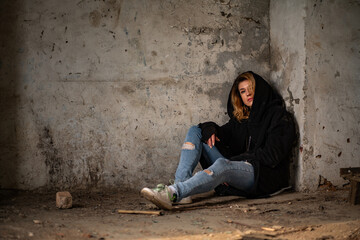 A mysterious goth woman in a black robe with a hood, torn jeans, sits thoughtfully on the dirty floor in an abandoned and destroyed building. Mysticism, post-apocalypse, depressed loneliness, witch - Powered by Adobe