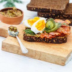 Delicious sandwich of rye bread with coriander with fried bacon, pickled cucumber, egg, dill and grainy mustard. A gourmet snack on a wooden board. Selective focus