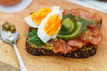 Delicious sandwich of rye bread with coriander with fried bacon, pickled cucumber, egg, dill and grainy mustard. A gourmet snack. Selective focus