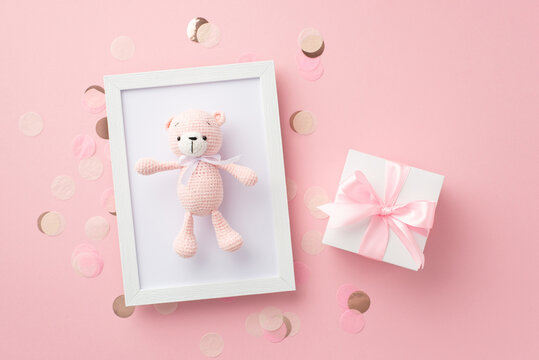 Baby girl concept. Top view photo of white giftbox with ribbon bow photo frame with knitted teddy-bear toy and shiny confetti on isolated pastel pink background