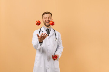 The male nutritionist doctor with stethoscope smiling and juggling tomatoes on beige background,...