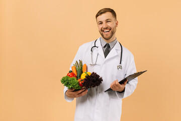 The happy male doctor nutritionist with stethoscope holding fresh vegetables on beige background,...