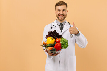 The happy male doctor nutritionist with stethoscope shows fresh vegetables on beige background,...