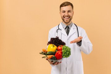 The happy male doctor nutritionist with stethoscope shows fresh vegetables on beige background, diet plan concept
