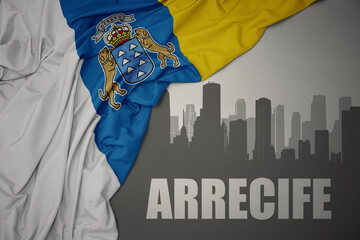 abstract silhouette of the city with text Arrecife near waving national flag of canary islands on a...