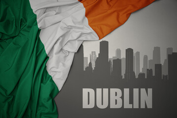 abstract silhouette of the city with text Dublin near waving national flag of ireland on a gray...