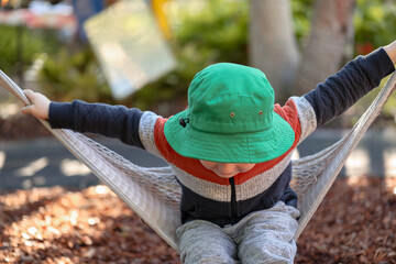 Little preschool boy playing on hammock at kindergarten with face hidden under bright green hat