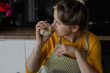 Closeup blond serious woman in striped apron smelling fresh garlic bulb in kitchen. Cooking detox...