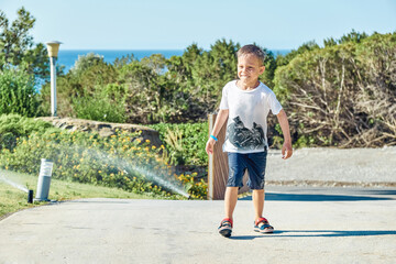 Preschooler boy wet through runs away from watering system on sunny day. Cute boy has fun on hotel...