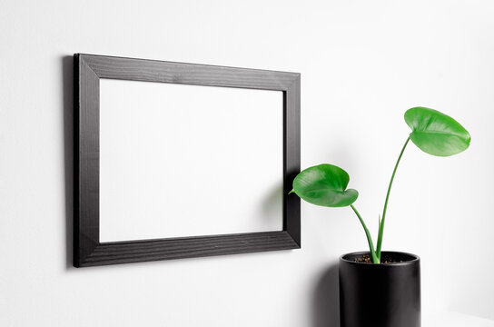 Black picture frame mockup over white wall and pot with home plant
