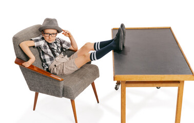 Cute little boy, happy kid in eyeglasses and hat sitting at armchair isolated on white studio background with copyspace for ad. Retro vintage style concept