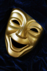 Portrait orientation image of gold comedy theatre mask surrounded by dark blue velvet fabric.