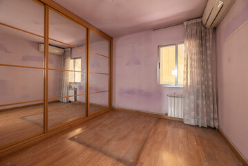 Empty bedroom with wardrobe with mirrored sliding doors, window with flower curtains, air conditioning and light oak parquet floor