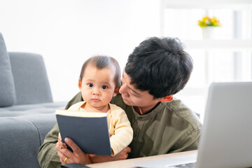 A baby daughter and an Asian father are reading a book together at home. and father has to work with