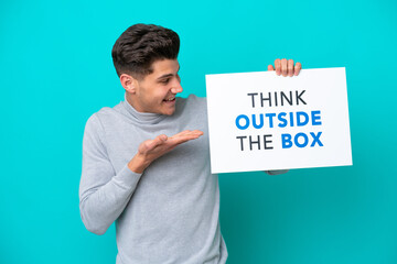 Young handsome caucasian man isolated on blue bakcground holding a placard with text Think Outside The Box and  pointing it