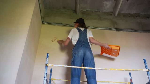 Female painter paints wall with roller on scaffolding at construction site. Young woman in overalls doing home decoration, renovation on staging. Professional worker makes DIY repair work in room.