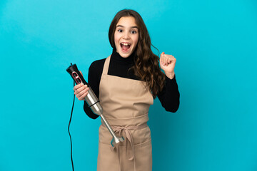 Little girl using hand blender isolated on blue background celebrating a victory in winner position