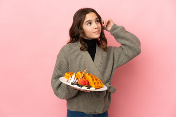 Little girl holding waffles isolated on pink background having doubts and thinking