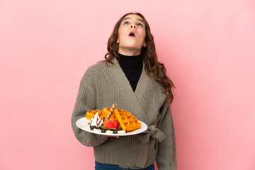 Little girl holding waffles isolated on pink background looking up and with surprised expression