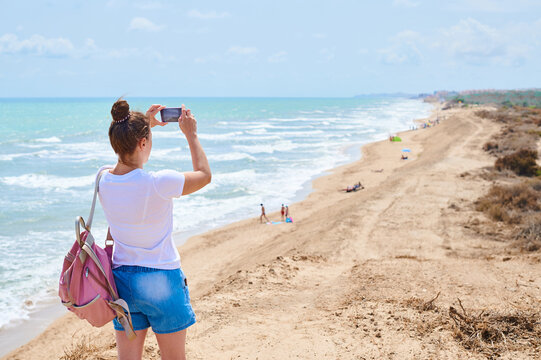 Young adult tourist woman with white shirt and jeans shorts takes pictures with her mobile from the top of a huge dune of a beautiful beach in Guardamar, Alicante, Spain, while enjoys summer vacation.
