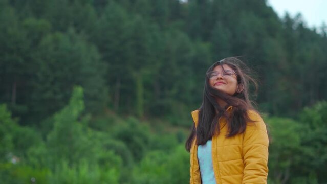 Portrait of happy Indian female standing in background of pine forest at Manali, Himachal Pradesh, India. Young girl with hair flying in the wind. Female enjoying nature during summer holidays 