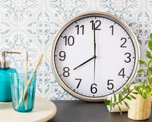 Fototapeta na wymiar Bathroom. Set with bottle of soap, glass with two teeth brush, clock and home plant on a marble table and tile wall. Cozy morning. Soft focus style image