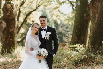 The bride and groom stand together between large old trees against the backdrop of a forest at...