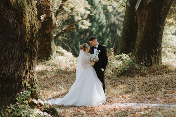 The bride and groom stand together between large old trees against the backdrop of a forest at...