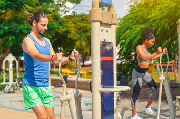 couple of afro dominican girl and white european boy exercising on bicycle machines in the park,...