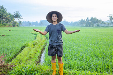 Naklejka na ściany i meble Attractive cheerful young Asian farmer standing, smiling and spreading arms showing success to growing paddy rice in the rice field. Modern agriculture concept. 