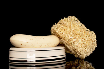 One loofah shower sponge and one bar of soap on a ceramic soap dish, close-up isolated on a black background.