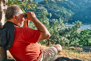 Tourist man resting in olive grove, looking through binoculars at the mountains view. Traveling or exercising outdoors. Active people, hiking, healthy lifestyle and harmony concept. Unity with nature.