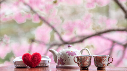 Soft Focus and Smooth Focus, a teapot and a set of ceramic crockery are placed on the table and there is a pink blurred background of the beautiful cherry blossoms blooming in winter.