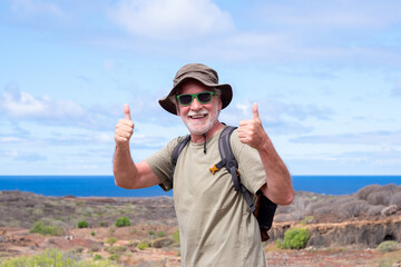 Happy active senior man with backpack and hat while hiking outdoors looking at camera with thumbs up. Blue sea and sky in the background