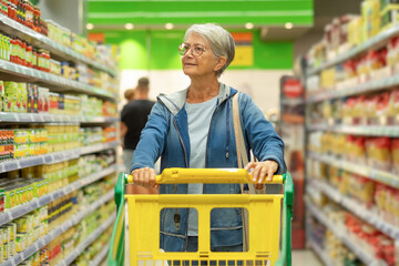 Caucasian senior woman pushing trolley in supermarket looking at the products on display, consumer...