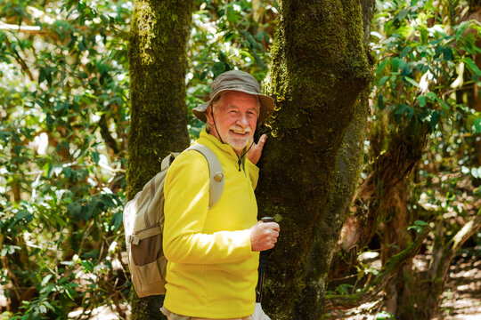 Cheerful senior bearded man with backpack and cap hiking in the forest enjoying healthy lifestyle and retirement,