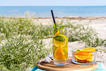 Detox infused water with lime, oranges, mint in glass on the beach with sea coast view