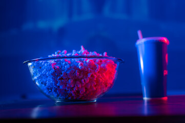 On a blue neon background, a glass bowl with popcorn, a drink in a blue glass. A traditional set for a cozy home stay near the TV. Watching your favorite movies and series, relaxing.