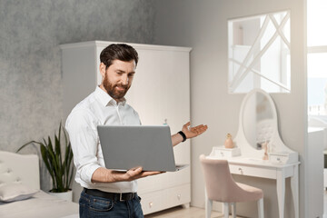Attractive male retailer demonstrating modern design of bedroom, while having online meeting remotely. Front view of bearded salesman using laptop for presenting showroom. Concept of interior design.