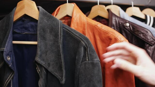 Man choosing clothing in a second hand store. Various vintage suede leather and jeans jackets hang on clothing rack. Thrifting and sustainability in clothing concept