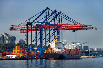 cargo ship moored for loading at container yard with container handling gantry crane or ship-to-shore crane unloading goods from deck to cargo port area, background cityscape with high-rise building.