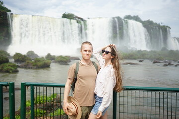In the photo, a beautiful girl and a guy stand against the backdrop of Iguazu Waterfalls located on the border of Brazil (Paraná state) and Argentina (Misiones province).