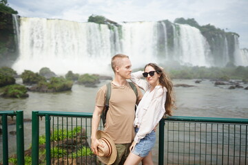 In the photo, a beautiful girl and a guy stand against the backdrop of Iguazu Waterfalls located on...