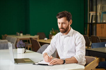 Confident, busy male manager making notes, while sitting at table in restaurant. Portrait of elegant entrepreneur in smart casual, working remotely with laptop indoors. Concept of working place.