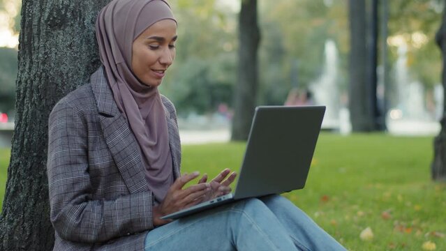 Happy smiling friendly islamic business woman muslim student girl teacher wearing hijab sitting on grass lawn in park near tree talking video call online chat conference waving hand greeting on laptop