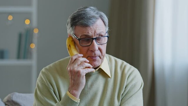 Dementia crazy elderly grandfather with glasses old man with psychological problems of schizophrenia funny senior male speaks in banana like phone pretending to call at home using fruit to calling