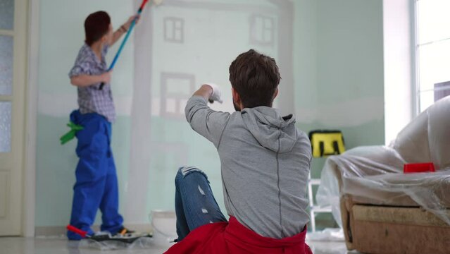 Back view man gesturing talking to blurred woman painting wall in new home. Joyful Caucasian young husband chatting with wife drawing house with paint roller at background. Renovation and family