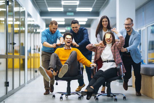 Young cheerful business people racing on office chairs and laughing. Funny activity, celebrating corporate success. Business concept.